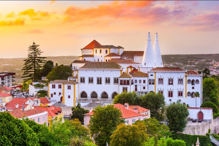 Stunning view of the National Palace of Sintra at sunset, with its iconic twin chimneys and whitewashed façade standing out above the red rooftops of the historic town. A cultural landmark and must-see stop on the Yes, You Deserve tour.