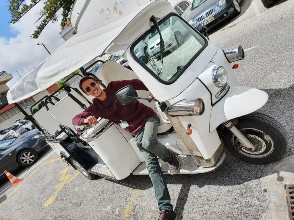 Daniel Ponce, smiling and wearing sunglasses, poses confidently in the driverโs seat of a white electric tuk-tuk during a Yes, You Deserve tour in Sintra, ready to lead another unforgettable adventure.