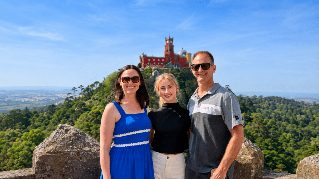 Three friends posing and smiling in front of Pena Palace atop a hill with blue sky and green trees around them by a rocky foreground.
