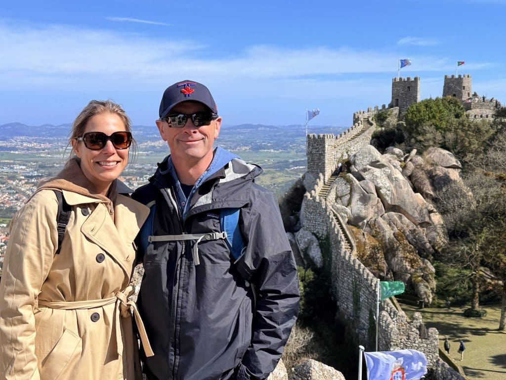 Smiling couple in jackets posing in front of a medieval stone castle on a hill under a clear blue sky.