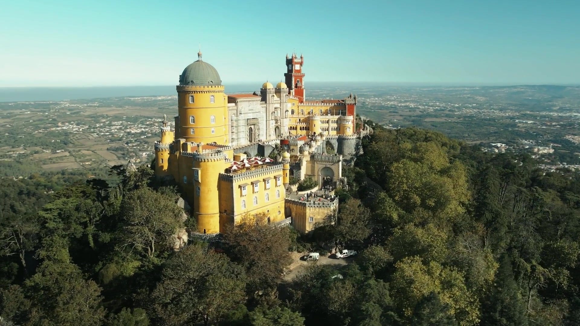 Pena Palace atop a forested hill with yellow walls and green dome, overlooking a valley and distant coastline.
