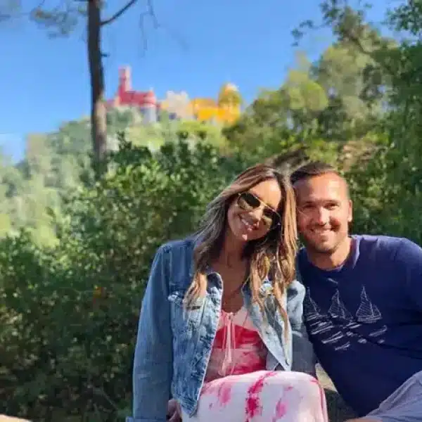 Smiling couple with Pena Palace in the background, enjoying a romantic tuk tuk tour in Sintra surrounded by lush greenery.