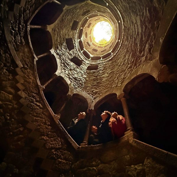 Spiral stone staircase winding upward in a tower, viewed from below with people looking toward the bright opening at the top.