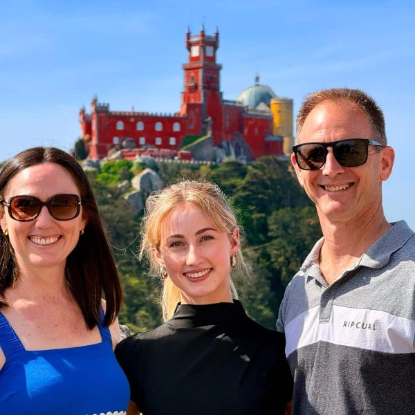 Three adults smiling for a photo outdoors with a bright red fortress on a hill behind them.