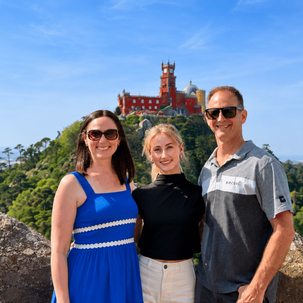 Three friends posing and smiling in front of Pena Palace atop a hill with blue sky and green trees around them by a rocky foreground.