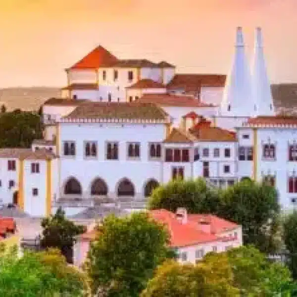 Sunset view of the National Palace of Sintra with its iconic twin chimneys, surrounded by colorful rooftops and lush greenery in the historic center of Sintra, Portugal.
